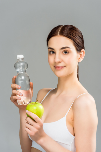 cheerful brunette girl holding bottle with water and green apple isolated on grey  - Photo, Image