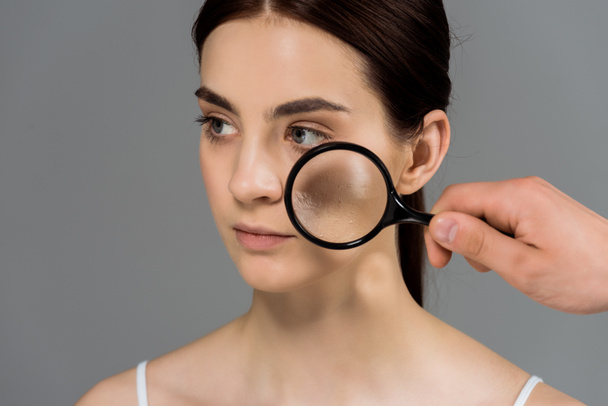 cropped view of man holding magnifier glass near woman with problem skin isolated on grey  - Foto, imagen