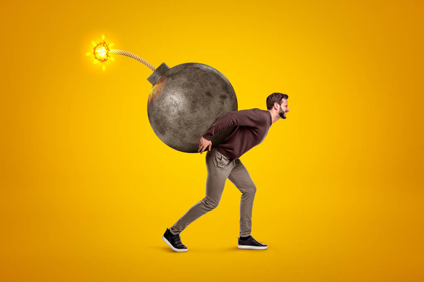 Young man in casual clothes carrying big ball bomb with fuse on his back on yellow background - Fotografie, Obrázek