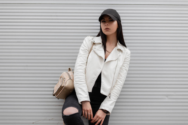 Young urban woman in a trendy white leather jacket in vintage ripped jeans in a stylish black baseball cap with a gold fashionable backpack stands near a silver metal wall. Beautiful girl. - Photo, Image