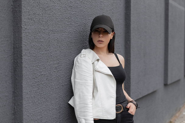 Beautiful pretty young woman in a stylish black baseball cap in a fashionable white leather jacket in vintage jeans in a t-shirt posing standing near a gray building in the city. Urban girl outdoors. - Photo, Image