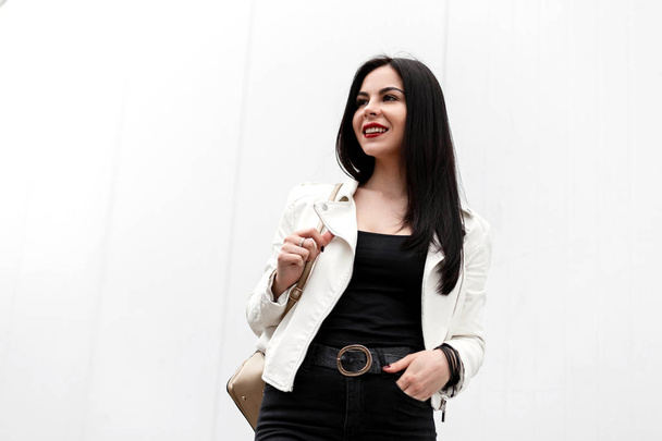 Hermosa joven morena con labios rojos en una sonrisa atractiva en una chaqueta blanca de moda en una camiseta con una mochila en jeans se relaja cerca de una pared blanca en la calle. Linda chica feliz
. - Foto, Imagen