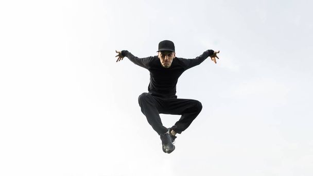 Młody sportowiec w czarnej koszuli w czapce w dżinsach w tenisówkach pozujący w skoku na tle błękitne niebo. Przystojny tancerz skaczący w powietrzu. - Zdjęcie, obraz