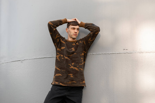 Young handsome man with a fashionable hairstyle in a stylish camouflage shirt in black vintage jeans is resting near a metallic gray wall. Attractive guy is relaxing outdoors on a sunny summer day. - Zdjęcie, obraz