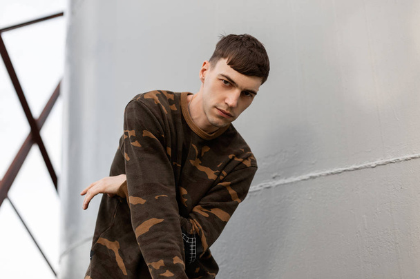 Stijlvolle knappe jonge man in een modieuze camouflage shirt met een trendy kapsel poses in de buurt van de grijze metalen wand buitenshuis. Aantrekkelijke kerel model. Herenkleding. - Foto, afbeelding
