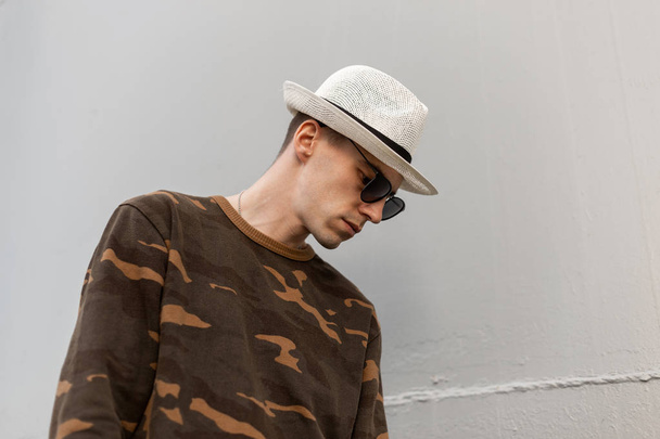 Porträt eines hübschen jungen Hipster-Mannes mit modischem Hut und stylischer dunkler Sonnenbrille im Militärhemd vor einer grauen Metallwand. American Modern Guy Mode-Modell. Seitenansicht. - Foto, Bild