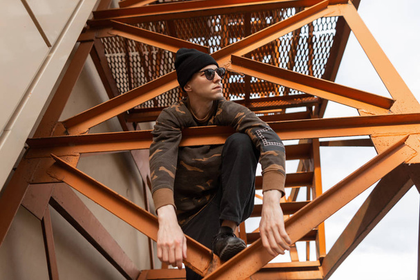 American young hipster man in a fashionable hat in black sunglasses in a vintage military shirt in jeans in a sneakers is sitting on the orange iron stairs outdoors. Nice guy is resting in the street. - Zdjęcie, obraz