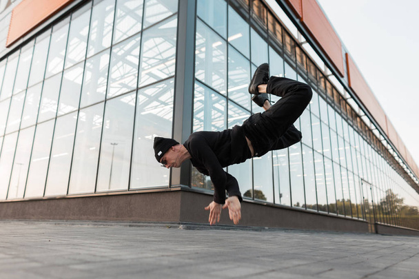 Athletic young man in black clothes in a stylish hat poses in the air outdoors near a glass modern building. Stylish dancer guy jumping. - Foto, Bild