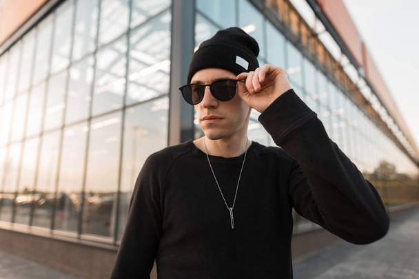 Young nice american hipster man in black shirt in knitted hat in fashionable sunglasses is posing standing near the glass building on the street. Handsome guy walks through the city on a summer day. - Foto, Imagen