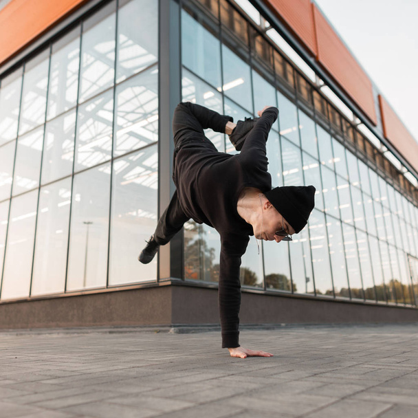 Nice young professional man dancer in black stylish clothes in a knitted hat in stylish sunglasses is doing a handstand near a glass modern building. Cool hipster guy dancing outdoors. Street style. - Photo, Image