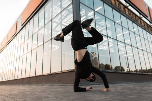 American handsome young hipster man in stylish dark sunglasses in trendy black clothes dancing break dance near a glass modern building in the city. Handsome professional guy dancer. - Photo, Image