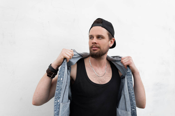 American young man hipster in a stylish black cap in a trendy T-shirt stands near a white wall in the city and straightens a fashionable denim vest. Handsome modern guy model posing. Street style. - Photo, Image