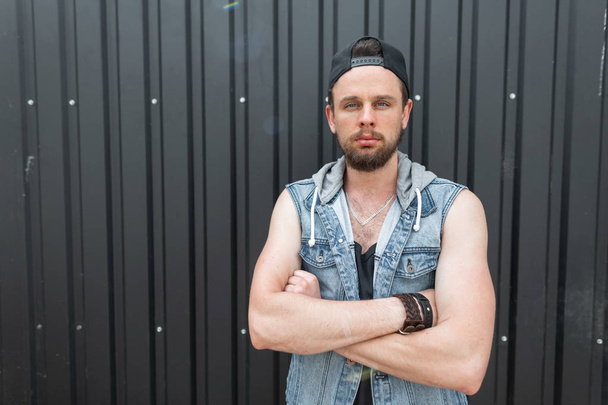 Handsome young hipster man in a fashionable blue denim vest in a t-shirt with a beard in a black stylish cap poses near metal black wall. Nice urban guy model outdoors. Modern men's street fashion. - Photo, Image