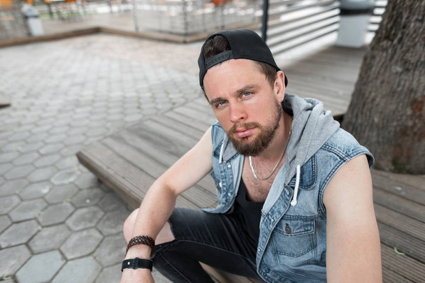 An einem warmen Sommertag sitzt ein junger Hipster mit Bart und T-Shirt in zerrissenen Jeans in einer schicken blauen Jeans-Weste und modischer Mütze auf einer Holzbank in der Stadt. amerikanischer gutaussehender Kerl - Foto, Bild