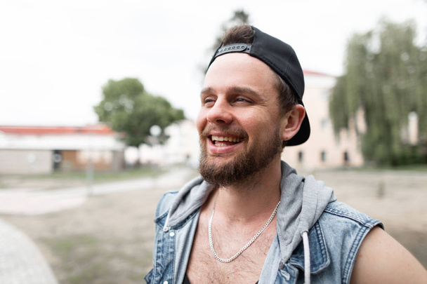 Positive young hipster man in a fashionable jeans vest in a stylish cap with a beard on the street on a warm summer day. Cheerful guy with a funny smile outdoors. - Photo, Image