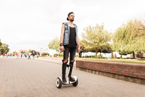 Young handsome man in ripped black jeans in a denim vest in a cap in a t-shirt in sneakers rides a hoverboard through the city on a summer sunny day. Urban guy on the white board. Leisure.  - Photo, Image