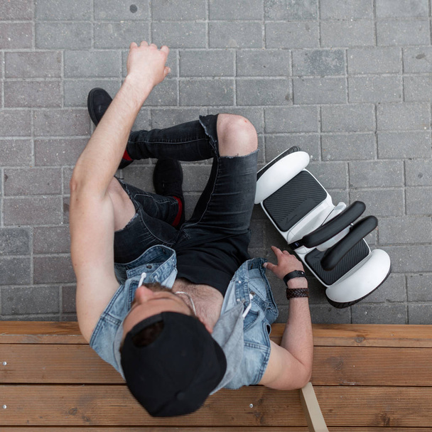 Guapo joven deportivo sentado en un escalón de madera junto a un moderno hoverboard blanco. La vista superior sobre el muchacho a la moda en la ropa de estilo en la gorra negra. Fin de semana activo saludable
. - Foto, Imagen