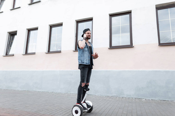 Young handsome man in ripped black jeans in a denim vest in a cap in a t-shirt in sneakers on a hoverboard around the city and talking on the phone on a sunny summer day. Urban guy on the white board. - Photo, Image