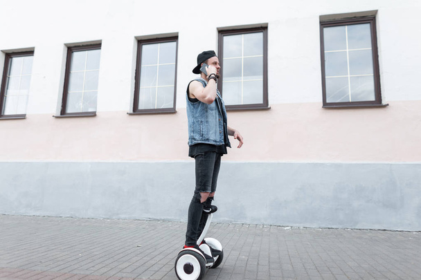 Attractive young man in a jeans vest in ripped jeans in a cap in a t-shirt in sneakers rides on a white gyroscope and talks on a mobile phone.Stylish guy travels around the city standing on hoveybord. - Photo, Image