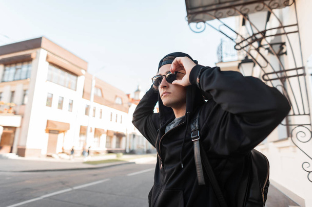 Stylish young hipster man in a trendy sweatshirt with a hood in dark sunglasses in a black cap with a stylish backpack travels around the city on a sunny summer day. Attractive guy outdoors. Menswear. - Foto, Bild