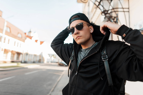 Attractive young man hipster in a stylish black hooded sweatshirt in a sunglasses in a trendy cap standing next a road near a vintage building in the city on a bright sunny day. Urban guy outdoors. - Фото, зображення