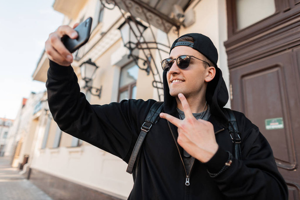 Stylish cheerful young man in a black sweatshirt with a hood in stylish sunglasses in a cap is smiling and making a selfie. Cheerful handsome hipster guy takes a picture and shows peace sign in camera - Foto, afbeelding