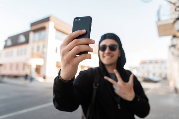Χαρούμενο νεαρό χίπστερ με κουκούλα σε γυαλιά ηλίου στο μοντέρνο καπέλο του μπέιζμπολ χαμογελάει και δείχνει την υπογραφή της ειρήνης κάνοντας selfie στο τηλέφωνο. Θετικός όμορφος τύπος περπατάει στο δρόμο κοντά σε vintage κτίρια. - Φωτογραφία, εικόνα