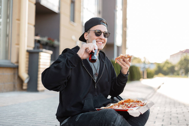 Счастливый молодой красивый мужчина в черной стильной одежде в кепке в солнечных очках сидит на улице и ест горячую пиццу и пьет содовую. Радостный американец обедает на улице. Время завтрака
. - Фото, изображение