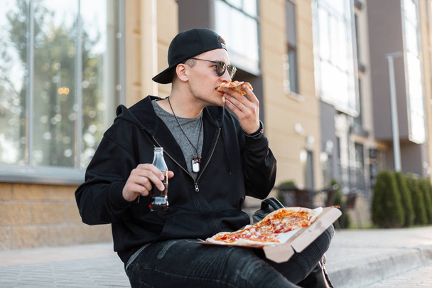 Urban joven guapo en negro ropa elegante en una gorra con gafas oscuras está sentado en la calle comiendo pizza caliente deliciosa y beber refrescos. Hambriento americano chico está almorzando al aire libre
.  - Foto, Imagen