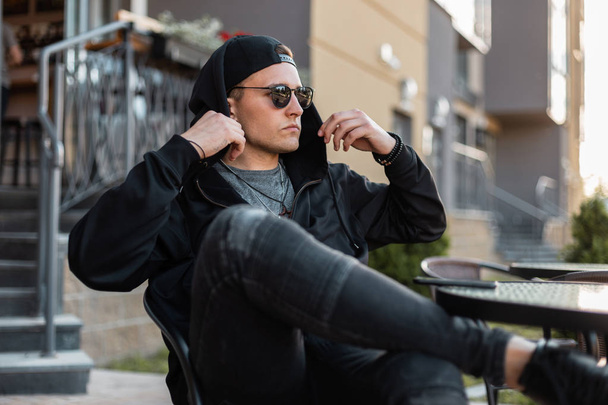 Mooie stedelijke jonge hipster man in zwarte hoodie in stijlvolle zonnebril in Vintage jeans in een zwarte pet rusten zitten in een zomercafé op een zomerdag. Knappe Amerikaanse kerel is ontspannen buitenshuis. - Foto, afbeelding