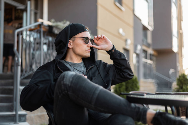 Amerikaanse jonge hipster man in stijlvolle zwarte kleding in modieuze zonnebril in een baseballpet in jeans zit in een zomercafé in de straat op een zonnige dag in de zomer. Knappe kerel rust buitenshuis. - Foto, afbeelding