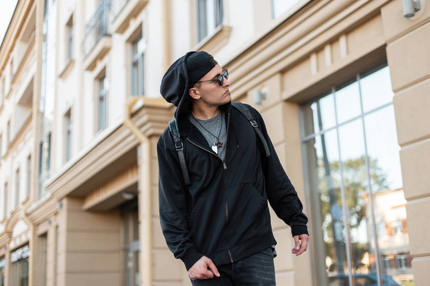 Handsome young hipster man in a hooded sweatshirt with a baseball cap in sunglasses with a backpack in jeans walking near a vintage building on a summer day. American guy traveling on the street. - Photo, Image