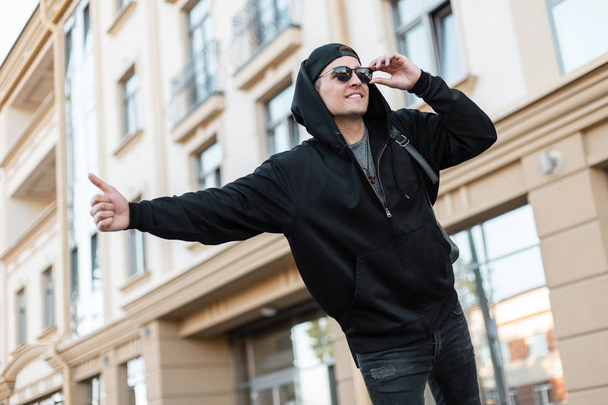 Fiatal elegáns hipster férfi divatos fekete ruhák napszemüveg sapka áll a városban, és fog egy taxit. Vonzó pozitív srác turista vár egy taxi. - Fotó, kép
