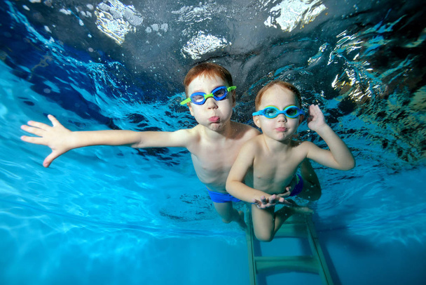 Twee vrolijke sport jongetjes zwemmen in het zwembad, op een blauwe achtergrond. Uitzicht vanaf de onderkant van het zwembad. Poseren voor de camera met je ogen open. Portret. Onderwater fotografie. Horizontale weergave - Foto, afbeelding