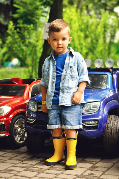 Um menino bonito de três anos fica perto de um carro elétrico preto em um parque com botas de borracha amarela e uma camisa jeans azul
. - Foto, Imagem