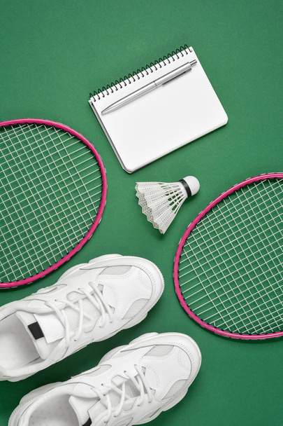 Sports equipment. Shuttlecock and badminton racket, sneakers, pen and notepad on green background. Fitness and healthy lifestyle concept. Flat lay, top view, copy space - Фото, изображение
