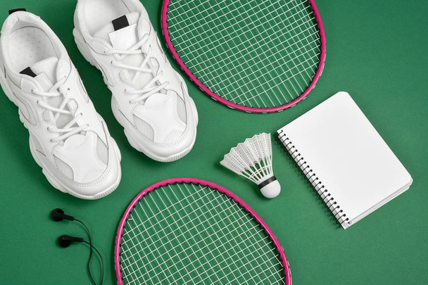 Sports equipment. Shuttlecock and badminton racket, sneakers, headphone, pen and notepad on green background. Fitness and healthy lifestyle concept. Flat lay, top view, copy space - Foto, afbeelding