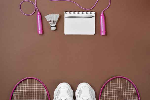 Sports equipment. Shuttlecock and badminton racket, skipping rope, sneakers, pen and notepad on brown background. Fitness and healthy lifestyle concept. Flat lay, top view, copy space - Foto, Bild