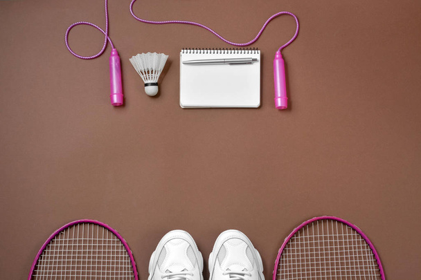 Sports flat lay with shuttlecock and badminton racket, skipping rope, sneakers, pen and notepad on brown background. Fitness, sport and healthy lifestyle concept. - Photo, image