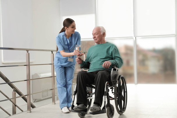 Nurse giving water to senior man in wheelchair at hospital. Medical assisting - Foto, Imagen