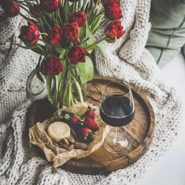 Wine snack set with flowers. Glass of red wine, cheese, roasted almonds, strawberries and bouquet of Spring red tulips on wooden tray over white knitted blanket, square crop. Romantic mood concept - Fotoğraf, Görsel