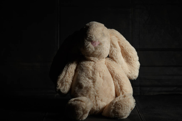 Miękka zabawka króliczka na brudnym ciemnym tle w wiązce twardego światła z cieniem. Koncepcja zaginionego dzieciństwa - Zdjęcie, obraz
