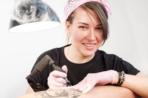 Female tattoo master tattooing female client. Figure nuns on the thigh. Black tattoo machine and lamp. Workplace artist. - Zdjęcie, obraz