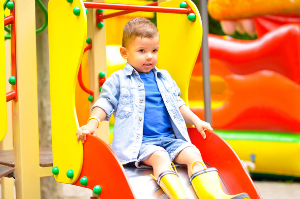 Pieni söpö kolmevuotias poika leikkii leikkikentällä ja liukuu alas liukumäkiä keltaisissa kumisaappaissa.
. - Valokuva, kuva