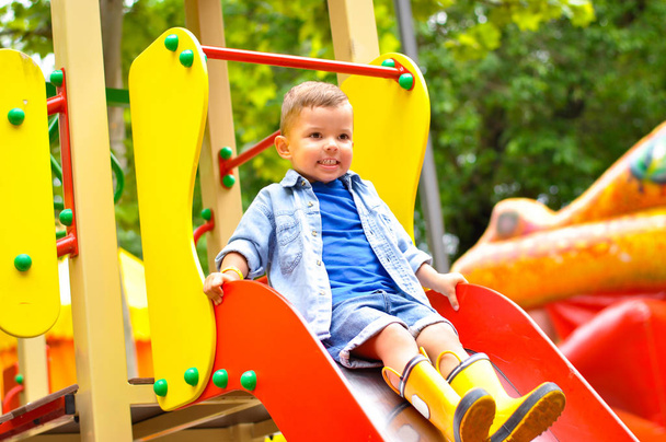 A little cute three-year-old boy plays on the playground and slides down the slides in yellow rubber boots. - Foto, imagen