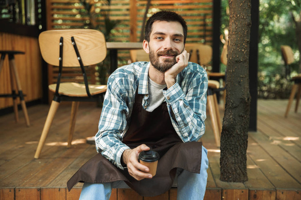 Photo of smiling waiter man sitting on wooden floor while workin - Zdjęcie, obraz