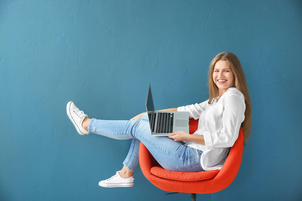 Beautiful young woman with laptop sitting on chair against color background - Photo, Image