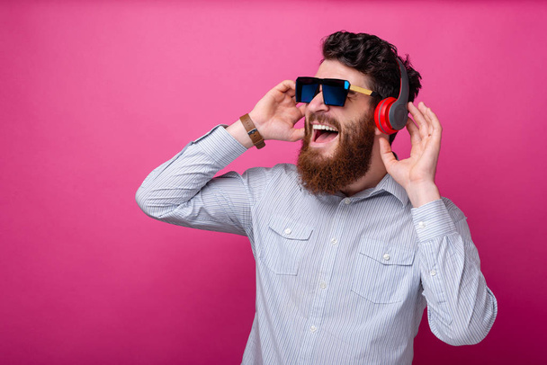 Homem barbudo bonito jovem ouvindo a música em fones de ouvido, vestindo óculos posando no fundo rosa
. - Foto, Imagem