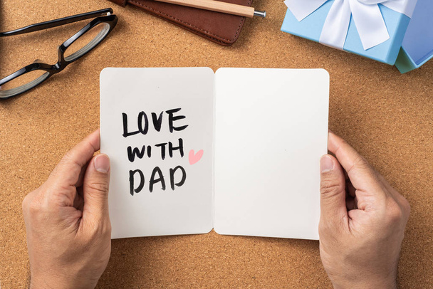 Fathers day layout with man hands holding white greeting card, ribbon kraft gift box on cork board background, copy space. - 写真・画像