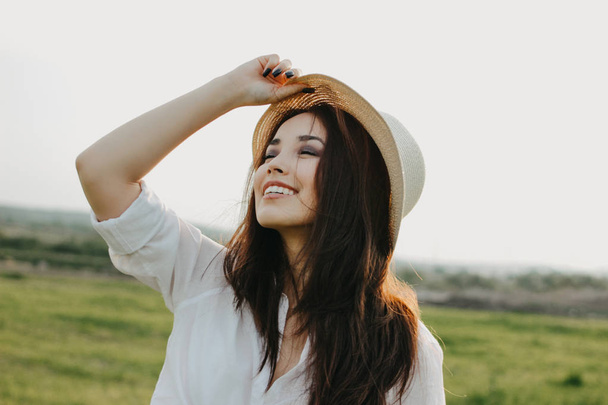 Belo cabelo longo despreocupado menina asiática em roupas brancas e chapéu de palha goza de vida no campo da natureza ao pôr do sol. Sensibilidade ao conceito de natureza
 - Foto, Imagem